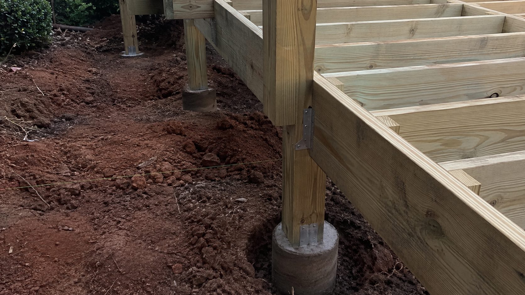 Deck post framing set on concrete footings in red clay soil, with joists ready for decking
